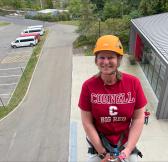 Amy in a red shirt and yellow helmet and rock climbing gear smiling up at the camera